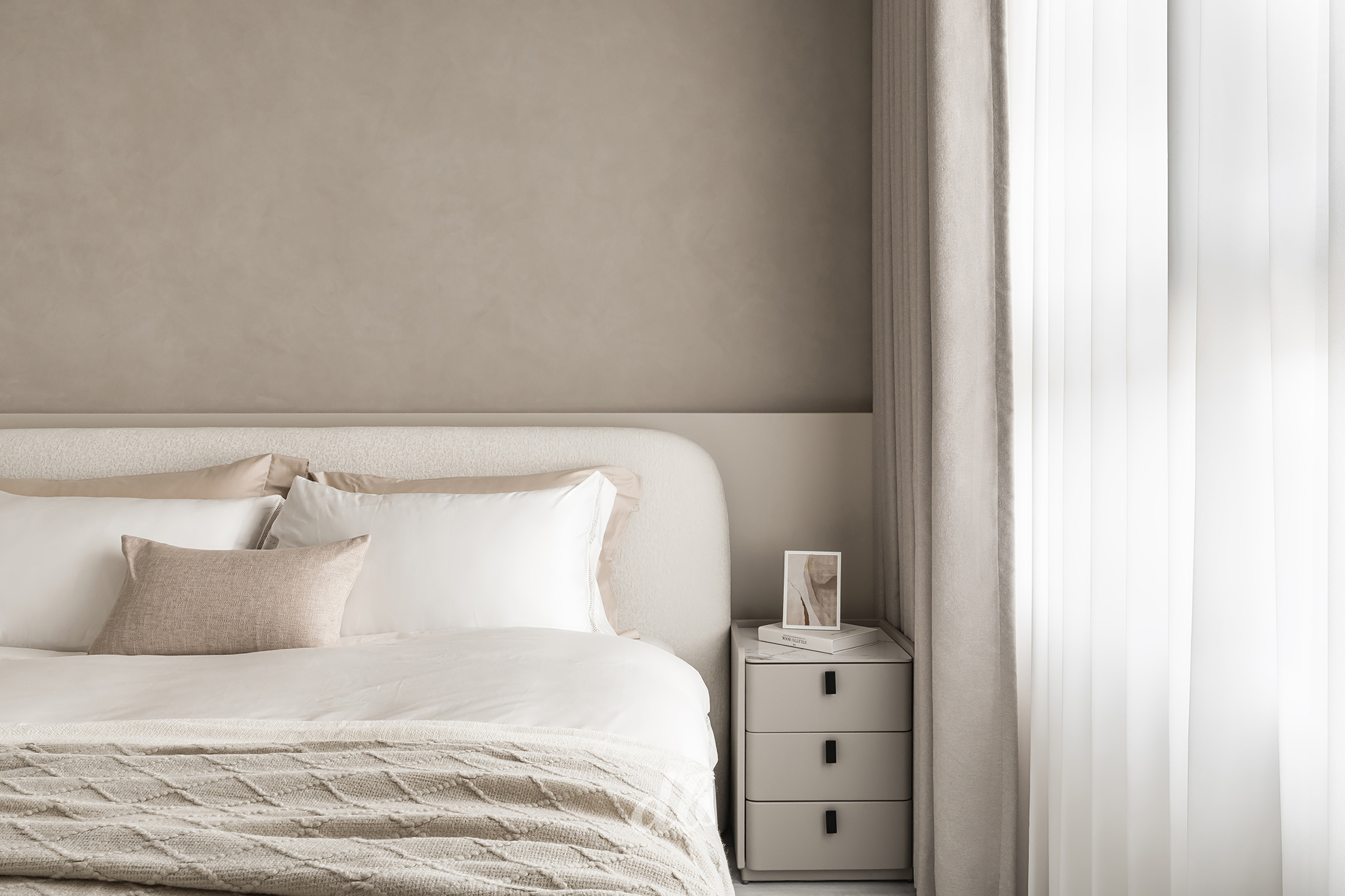 韓式奶油風臥室日造設計案例：選用高明度的暖灰色調牆面，搭配圓潤造型的布藝床頭板與米色系寢具，透過編織毯的異材質疊加與輕柔紗簾灑落的自然光影，營造出極簡、溫潤且具備舒壓質感的韓系睡眠空間。